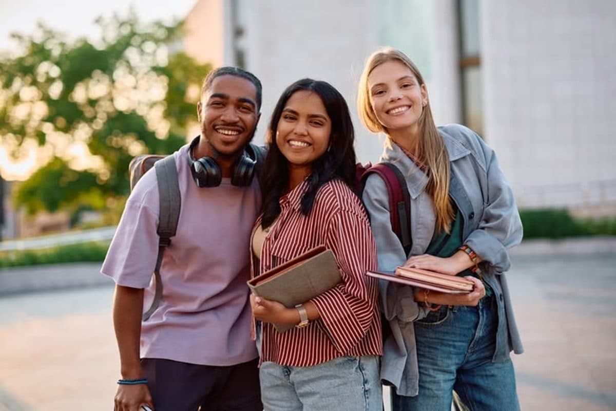 Three students smiling together on campus, carrying backpacks and notebooks