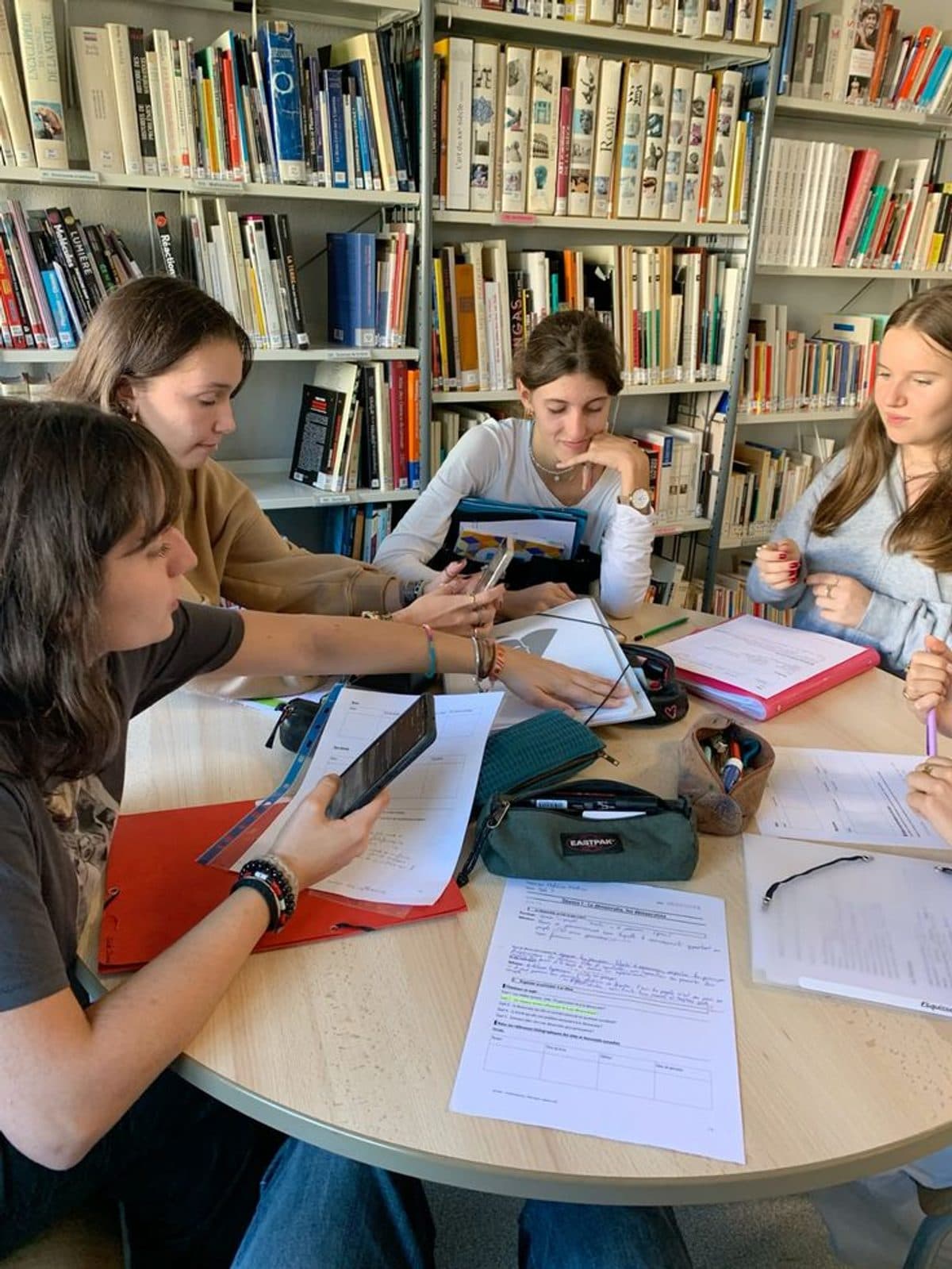 Four students studying together at a library table surrounded by books and notes